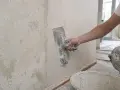 Ręka trzymająca kielnię wygładza tynk na ścianie. Widać starą, łuszczącą się farbę obok świeżo nałożonej gładzi.