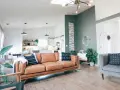 Nowoczesny salon z brązową kanapą i szarym fotelem. Ściana w kolorze zielonym dodaje charakteru, a rośliny wprowadzają życie. Idealny kolor do salonu.