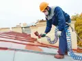 Pracownik na dachu maluje metalową dachówkę, używając pistoletu natryskowego. Wybiera odpowiednią farbę na rdzę, by zabezpieczyć konstrukcję.