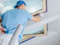 Malowanie poddasza to ważny etap wykończenia mieszkania krok po kroku. Pracownik zabezpiecza okna taśmą i przygotowuje się do malowania wałkiem.