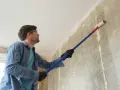 Mężczyzna w okularach ochronnych i rękawiczkach wykonuje gruntowanie ściany przed malowaniem, używając wałka na wysięgniku.