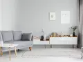 Sofa w kolorze popielatym, biały stolik z książką, komoda z dekoracjami i obrazki na ścianie.