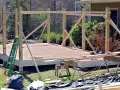 Drewniana konstrukcja altany w trakcie budowy. Widać gotowy taras, słupy i belki, drabinę oraz materiały budowlane. To pierwszy etap, jak zbudować altanę.