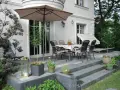 Uroczy leśny ogród z tarasem, stołem i krzesłami pod parasolem. Wokół bujna zieleń i kwiaty.