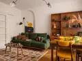 Zielona kanapa, drewniana biblioteczka i żółte krzesła tworzą przytulne wnętrze w stylu prl.