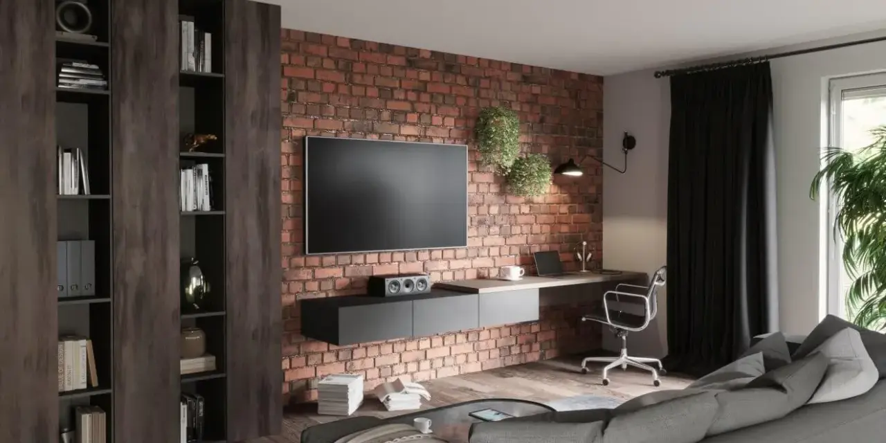 Nowoczesny salon loft inspiracje: ceglana ściana, wisząca szafka RTV, biurko z laptopem, regały i szara kanapa.