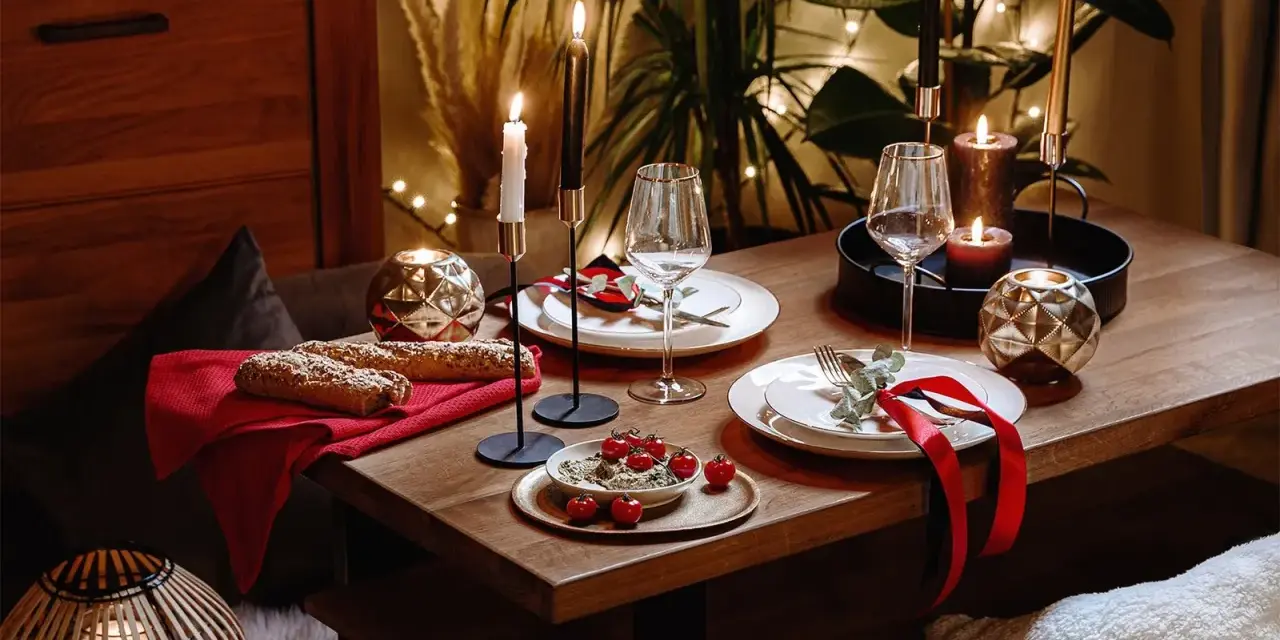 Romantyczna kolacja w domu: stół nakryty do świętowania, świece, kieliszki, pieczywo i pomidorki. Wystrój tworzy przytulną atmosferę.