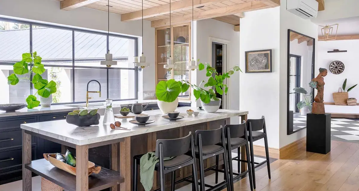 Nowoczesny salon w stylu modern farmhouse z wyspą kuchenną, drewnianymi belkami na suficie i dużą ilością zieleni.