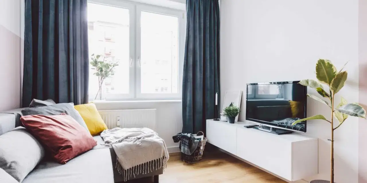 Przytulny salon w bloku z elementami feng shui: sofa z poduszkami, telewizor, roślina i zasłony tworzą harmonię.