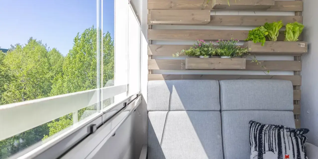 Przytulny mały balkon w bloku z szarą sofą, poduszką z liskiem i zielenią na drewnianej ściance.