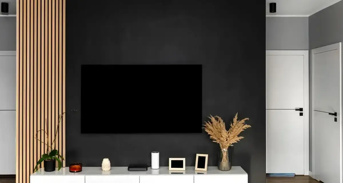 Nowoczesny salon z czarną ścianą, na której wisi telewizor. Obok drewniana ażurowa ścianka i biała szafka RTV.