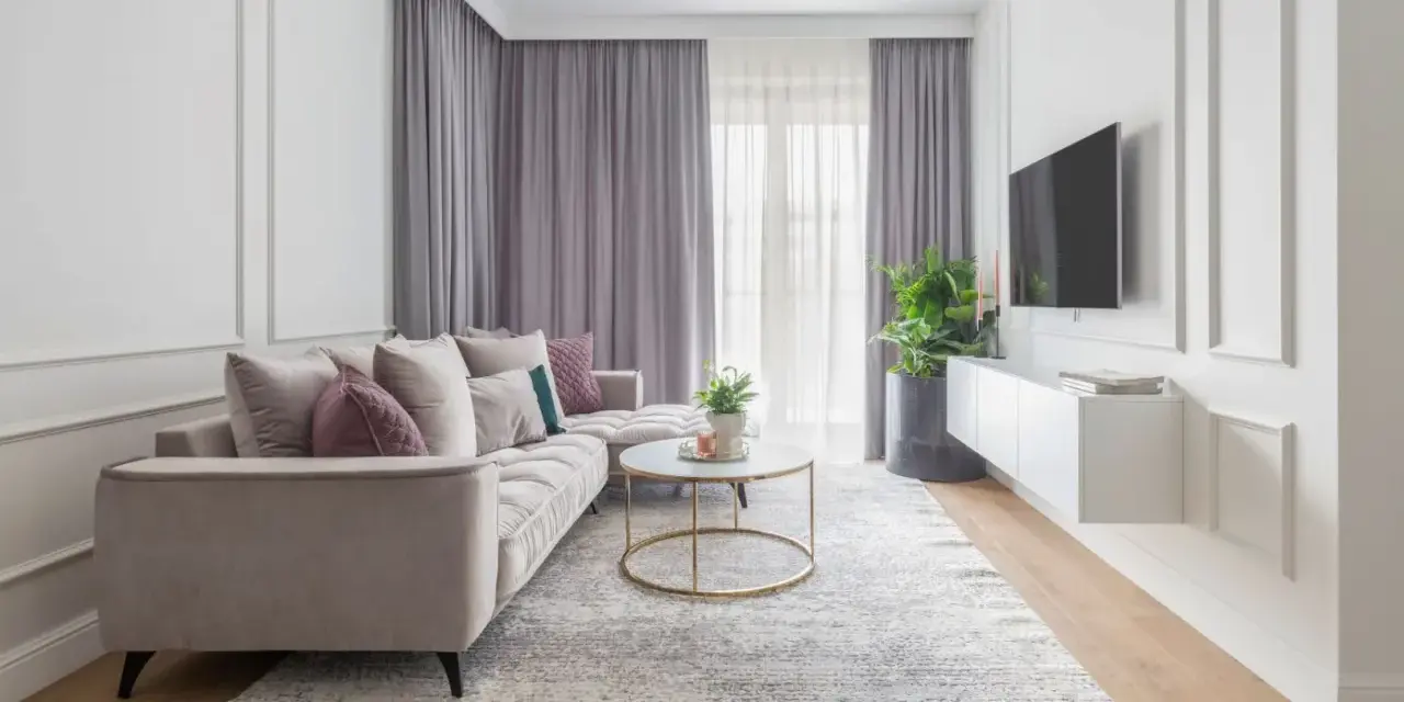 Elegancki salon w stylu modern classic. Jasna kanapa, okrągły stolik kawowy, dywan i zasłony tworzą przytulną atmosferę.
