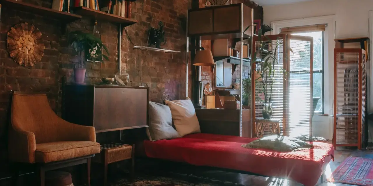 Przytulna sypialnia w stylu retro z ceglaną ścianą, regałami pełnymi książek, wygodnym fotelem i łóżkiem z czerwoną narzutą.