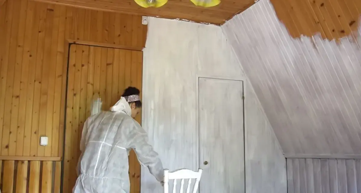 Osoba w kombinezonie maluje boazerię na biało. Widać postęp prac, część ścian jest już pomalowana.