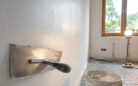 Nakładanie gładzi na ściany za pomocą kielni. W tle okno i wiadro z masą szpachlową.