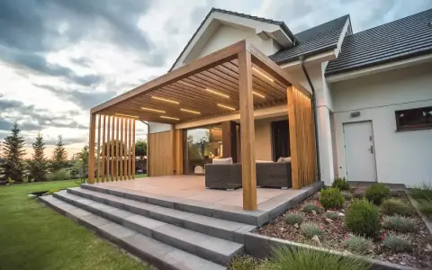 Nowoczesny taras z drewnianą pergolą i oświetleniem LED. Elegancka, jasnoszara jaka podłoga na taras, schody i meble wypoczynkowe tworzą spójną całość.