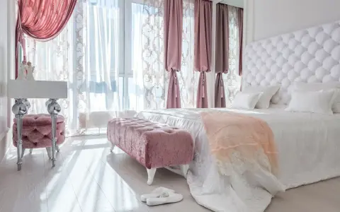 Elegancka sypialnia w stylu angielskim z pikowanym zagłówkiem, różowymi pufami i zasłonami.