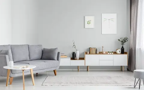 Sofa w kolorze popielatym, biały stolik z książką, komoda z dekoracjami i obrazki na ścianie.