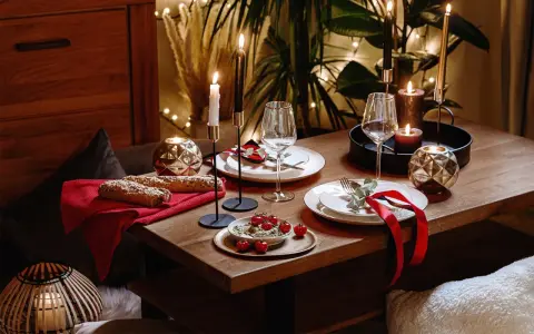 Romantyczna kolacja w domu: stół nakryty do świętowania, świece, kieliszki, pieczywo i pomidorki. Wystrój tworzy przytulną atmosferę.