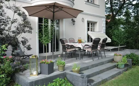 Uroczy leśny ogród z tarasem, stołem i krzesłami pod parasolem. Wokół bujna zieleń i kwiaty.