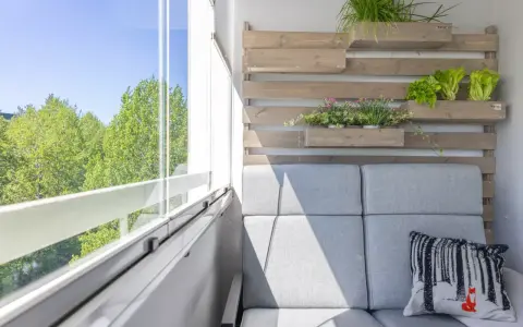 Przytulny mały balkon w bloku z szarą sofą, poduszką z liskiem i zielenią na drewnianej ściance.