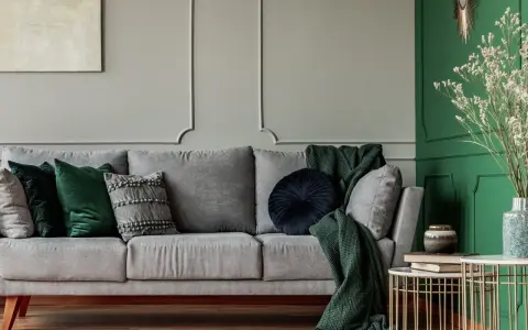 Szaro-zielony salon z wygodną sofą, ozdobioną poduszkami i kocami. Obok stoi stolik z książkami i wazonem.