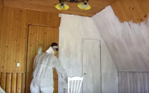 Osoba w kombinezonie maluje boazerię na biało. Widać postęp prac, część ścian jest już pomalowana.