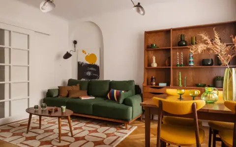 Zielona kanapa, drewniana biblioteczka i żółte krzesła tworzą przytulne wnętrze w stylu prl.