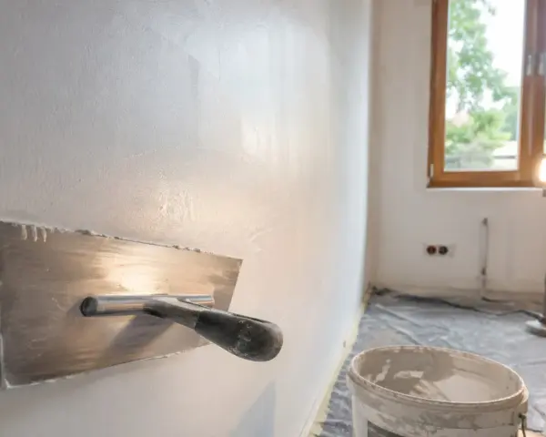 Nakładanie gładzi na ściany za pomocą kielni. W tle okno i wiadro z masą szpachlową.