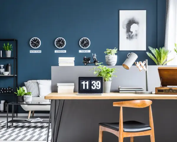 Biuro z zegarami pokazującymi czas w Nowym Jorku, Londynie i Tokio na ścianie w kolorze navy. Na biurku laptop, lampka i gramofon.