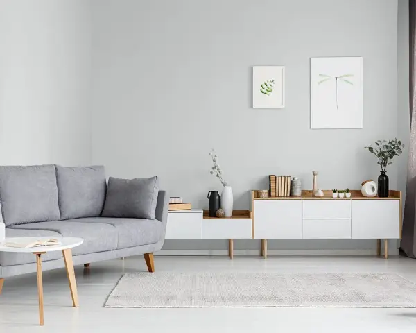 Sofa w kolorze popielatym, biały stolik z książką, komoda z dekoracjami i obrazki na ścianie.