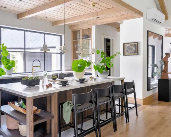Nowoczesny salon w stylu modern farmhouse z wyspą kuchenną, drewnianymi belkami na suficie i dużą ilością zieleni.