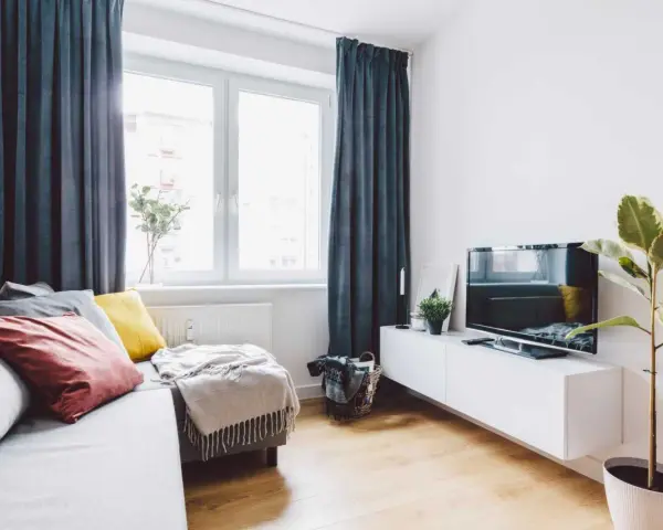 Przytulny salon w bloku z elementami feng shui: sofa z poduszkami, telewizor, roślina i zasłony tworzą harmonię.