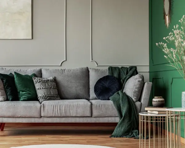 Szaro-zielony salon z wygodną sofą, ozdobioną poduszkami i kocami. Obok stoi stolik z książkami i wazonem.