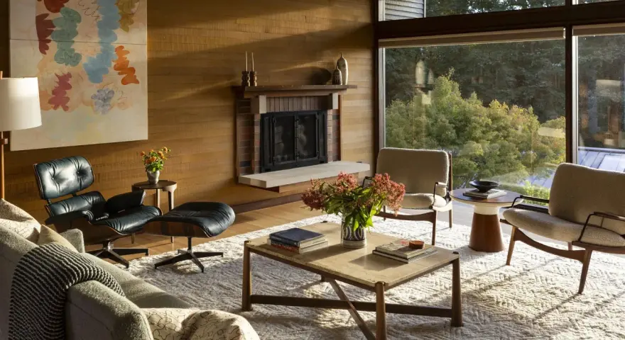 Salon w stylu mid century modern z kominkiem, wygodnymi fotelami i dużym oknem z widokiem na zieleń.