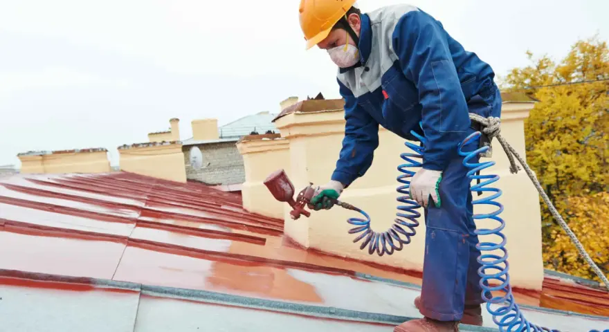 Pracownik na dachu maluje metalową dachówkę, używając pistoletu natryskowego. Wybiera odpowiednią farbę na rdzę, by zabezpieczyć konstrukcję.