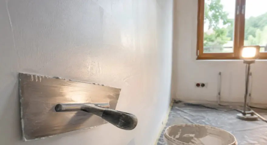 Nakładanie gładzi na ściany za pomocą kielni. W tle okno i wiadro z masą szpachlową.