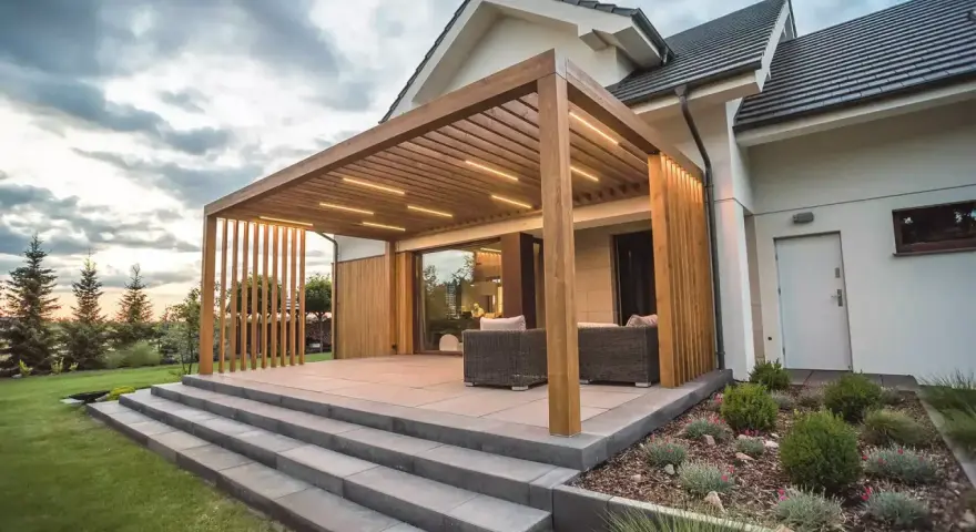 Nowoczesny taras z drewnianą pergolą i oświetleniem LED. Elegancka, jasnoszara jaka podłoga na taras, schody i meble wypoczynkowe tworzą spójną całość.