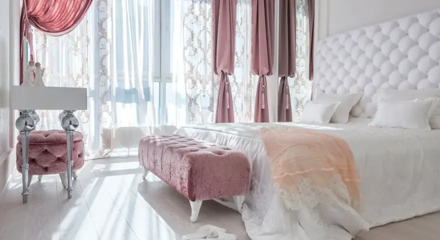 Elegancka sypialnia w stylu angielskim z pikowanym zagłówkiem, różowymi pufami i zasłonami.