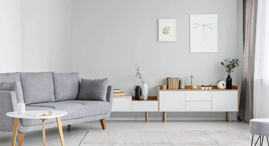 Sofa w kolorze popielatym, biały stolik z książką, komoda z dekoracjami i obrazki na ścianie.
