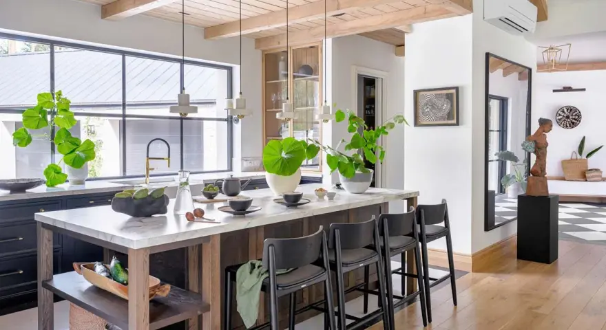 Nowoczesny salon w stylu modern farmhouse z wyspą kuchenną, drewnianymi belkami na suficie i dużą ilością zieleni.