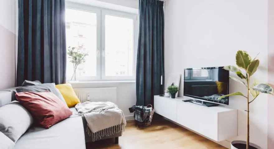 Przytulny salon w bloku z elementami feng shui: sofa z poduszkami, telewizor, roślina i zasłony tworzą harmonię.