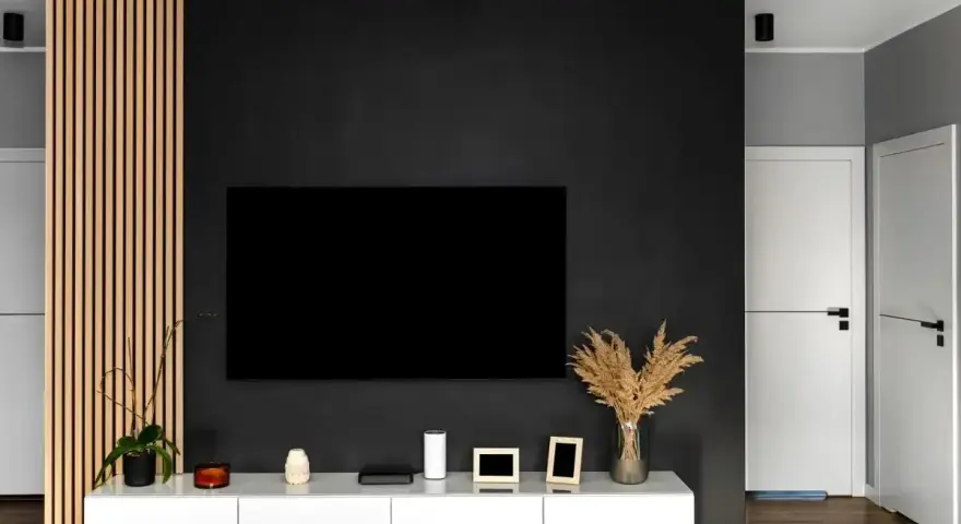 Nowoczesny salon z czarną ścianą, na której wisi telewizor. Obok drewniana ażurowa ścianka i biała szafka RTV.