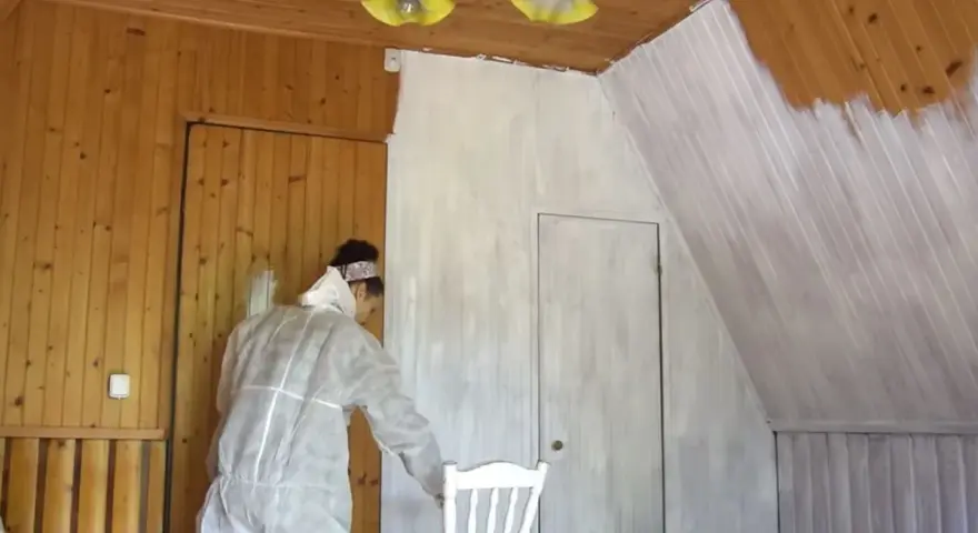 Osoba w kombinezonie maluje boazerię na biało. Widać postęp prac, część ścian jest już pomalowana.
