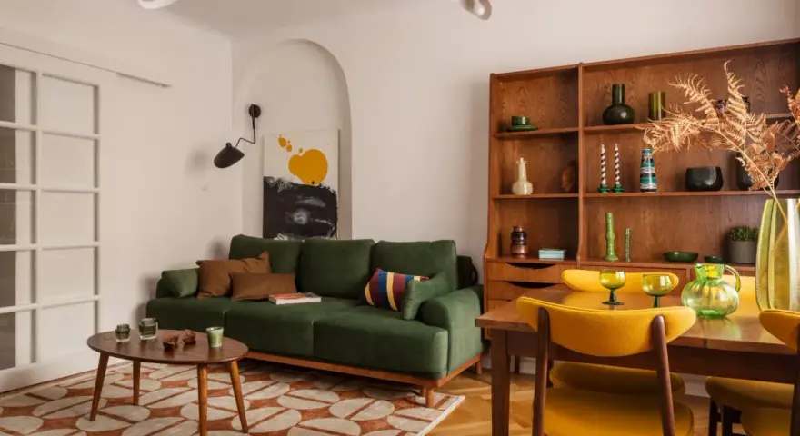 Zielona kanapa, drewniana biblioteczka i żółte krzesła tworzą przytulne wnętrze w stylu prl.