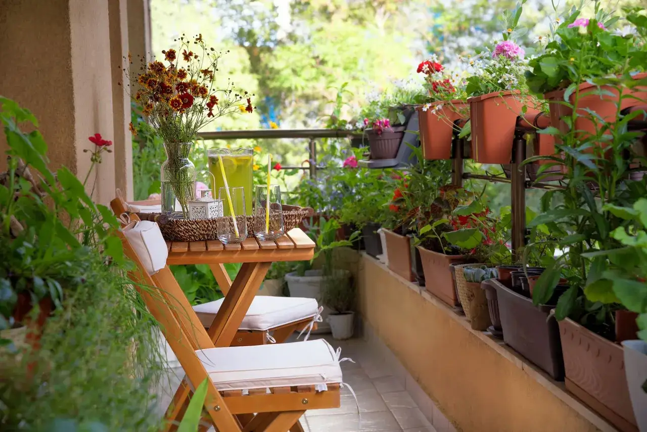 Przytulny balkon w bloku, pełen zieleni i kwiatów. Drewniany stolik z napojem i krzesła zapraszają do relaksu.