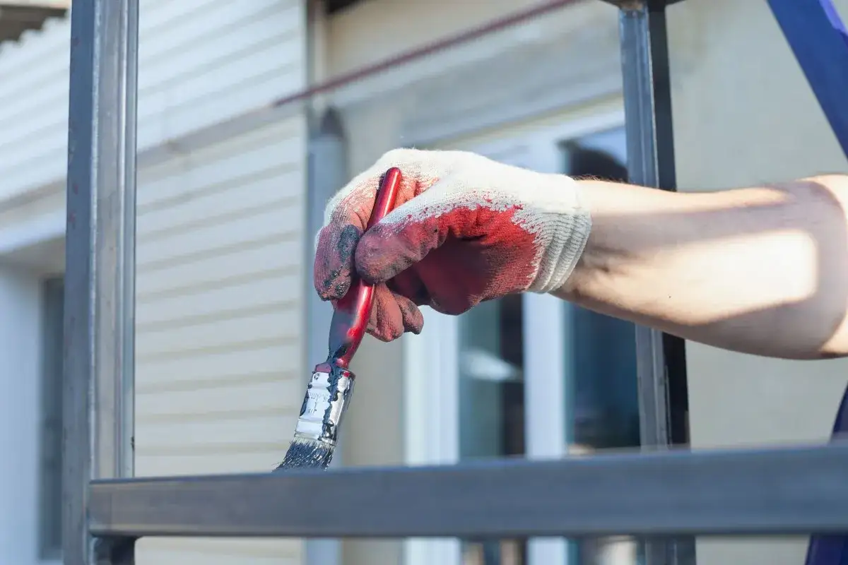 Ręka w rękawicy maluje pędzlem metalową balustradę. Szukasz odpowiedniej farby do metalu na zewnątrz?