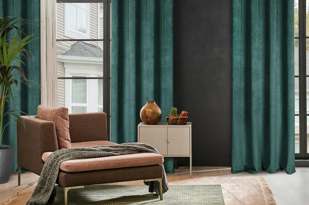 Eleganckie, ciemnozielone zasłony pomagają jak ochłodzić pok&oacute;j, tworząc przytulną atmosferę. Brązowa sofa z narzutą i rośliny dodają ciepła.