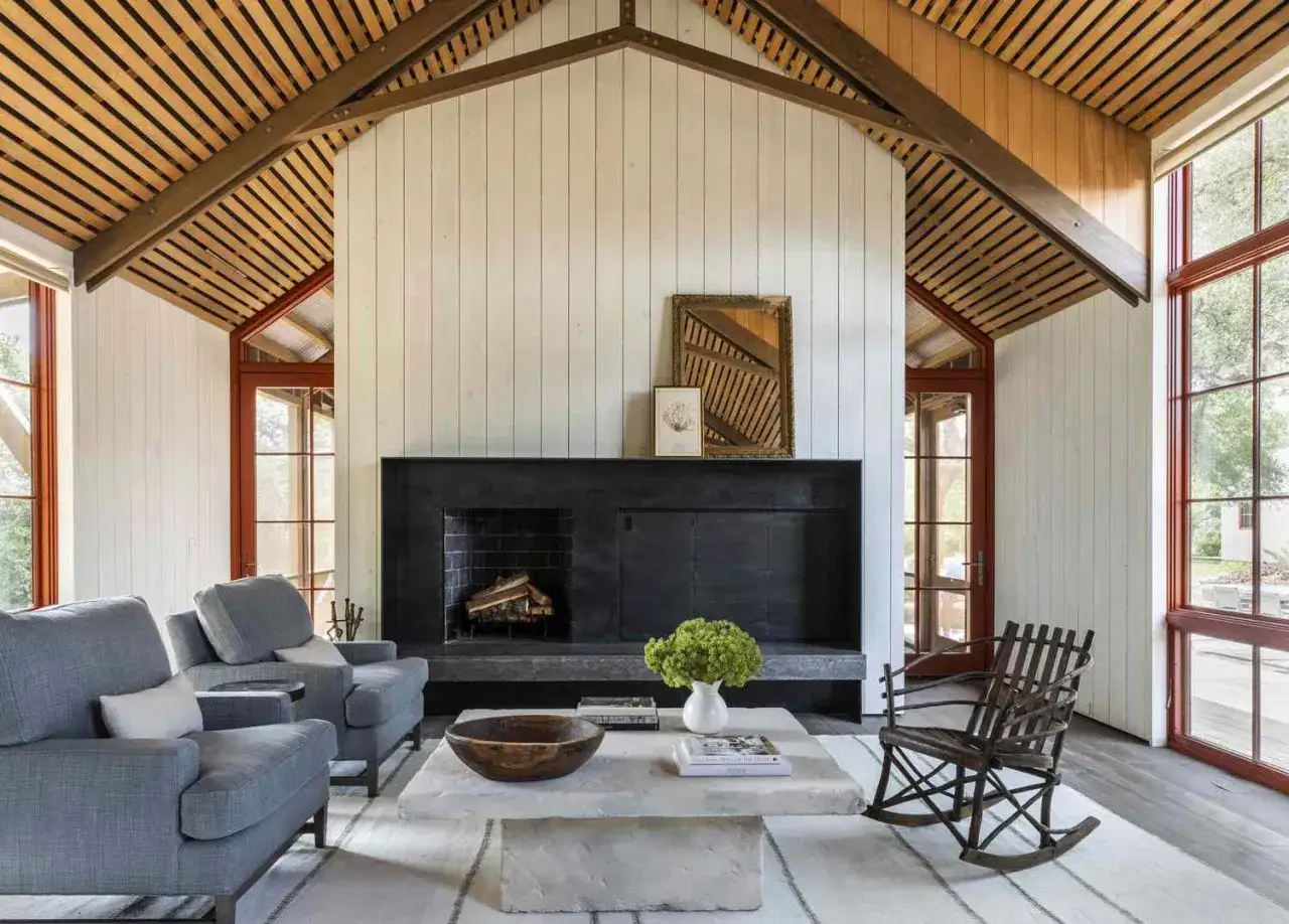 Przytulny salon w stylu modern farmhouse z kamiennym kominkiem, drewnianym sufitem i wygodnymi szarymi fotelami.