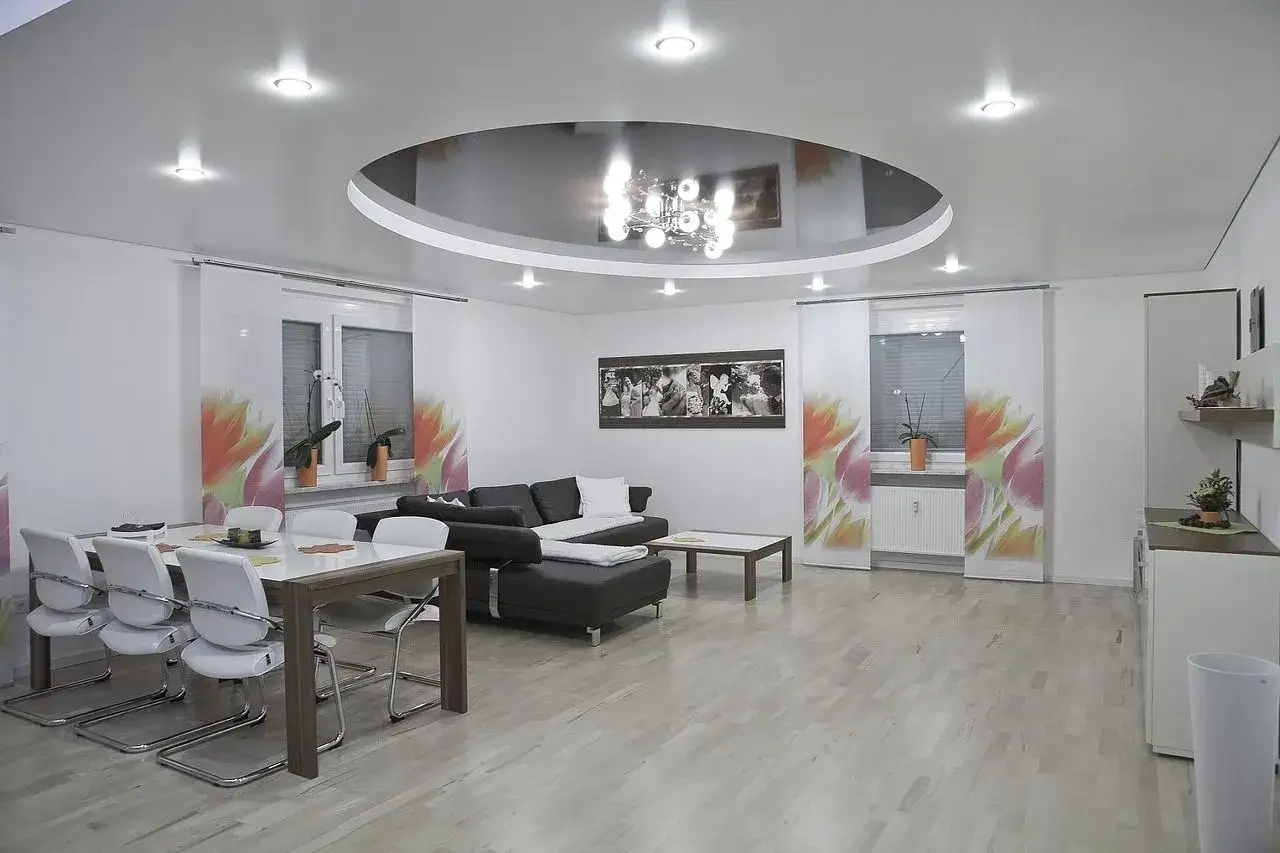Nowoczesny salon z okrągłym sufitem napinanym, który odbija elegancki żyrandol. Wnętrze zdobią białe meble i zasłony z motywem kwiatowym.